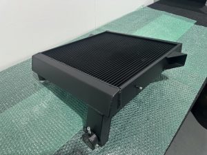 A black metal automotive radiator resting on green bubble wrap, positioned on a flat surface against a white wall.