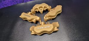 Four gold-colored Tokico brake calipers are arranged on a black surface, with the Tokico logo visible on two of them.