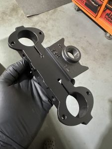 A gloved hand holds a black metal automotive part with circular openings, resting on a gray surface near an orange tool chest.