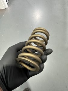 A gloved hand holds a metallic, gold-colored coil spring against a gray floor background.