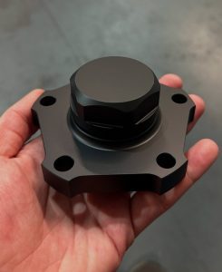 A hand holds a machined metal component with a hexagonal center and four mounting holes, set against a blurred gray background.