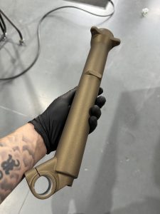 A gloved hand holds a metallic, gold-colored cylindrical component with a mounting hole, against a smooth gray floor background.