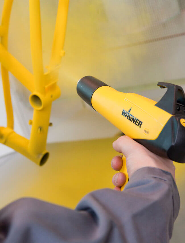 A person using a Wagner handheld paint sprayer for a PB powder coating application on a yellow bicycle frame.